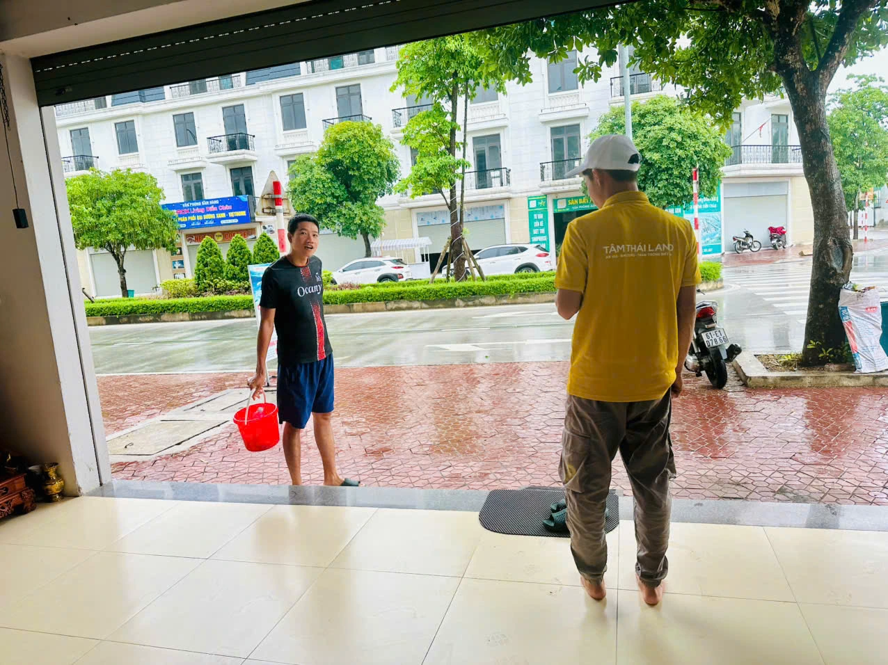 Hai người lớn trao đổi trước cửa; một người cầm xô đỏ, người còn lại mặc áo vàng.